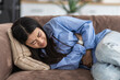 © kucherav - Young Asian woman suffering from abdominal pain lying on the couch at home. Female suffering from severe spasms holding her stomach