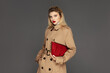 © Beauty Agent Studio - Fashionable young model posing against grey background dressed in trench coat and holding stylish red bag. Autumn fashion concept