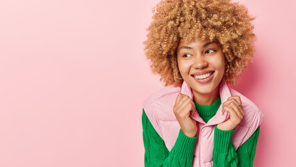 Wall Mural - Studio shot of beautiful woman smiles broadly shows white teeth wears green jumper and vest isolated over pink background blank space for your advertising content. Positive human emotions concept