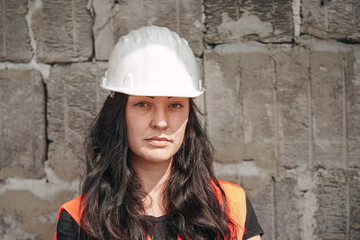 Young woman in white hard hat and orange high visibility vest, long dark hair, looking into camera. Blurred old construction site wall background