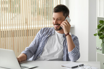 Poster - Happy man talking on smartphone while working with laptop in office