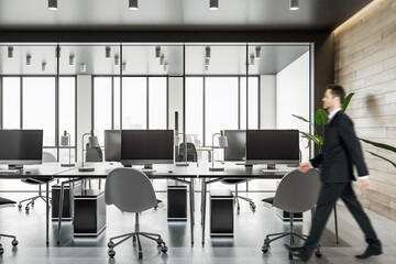 Canvas Print - Businessman walking by workspace tables with modern computers in spacious coworking office with wooden wall and panoramic window