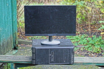 an old black desktop computer and a large dirty monitor stand on a gray wooden table on the street against the backdrop of green vegetation