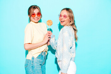 Wall Mural - Two young beautiful blond smiling hipster female in trendy summer clothes. Sexy carefree women posing near blue wall in studio. Trendy and positive models having fun with candy sweet lollipop