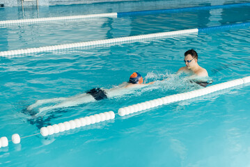 Wall Mural - A coach teaches a seven-year-old boy to swim in a modern swimming pool. Learn to swim. Development of children's sports. Healthy parenting and promotion of children's sports.