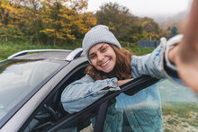 Young cheerful woman driver in a hat and jacket traveling by car taking a selfie on a smartphone while standing next to the car in the autumn forest