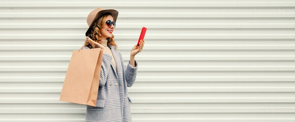 Wall Mural - Portrait of happy smiling young woman taking selfie with smartphone holding shopping bags wearing coat, round hat on white background, blank copy space for advertising text