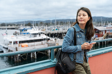 Canvas Print - smiling asian Korean woman visitor waiting for friend at railing with phone in hand at Old Fisherman's Wharf in California. a fleets of boats are staying in the fishing harbor at background