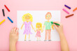 Baby girl hands drawing happy family with colorful wax crayons on light pink table background. Pastel color. Toddler development. Point of view shot. Closeup. Top down view.