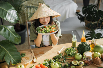 Joyful asian woman in traditional conical hat holding salad from organic vegetables and fruits enjoying healthy dinner in exotic light studio in tropical resort. Food and nutrition for weight loss.