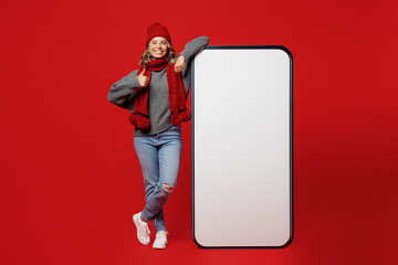 Wall Mural - Full body young woman wear grey sweater scarf hat big blank screen mobile cell phone with area show thumb up isolated on plain red background. Healthy lifestyle ill sick disease cold season concept.