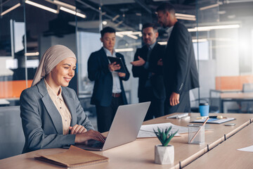 Wall Mural - Smiling muslim businesswoman in hijab working on computer while sitting in modern office