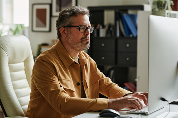 Wall Mural - Mature businessman with grey hair and beard typing on keyboard while sitting by workplace in front of computer monitor in office