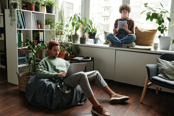 Wall Mural - Young woman with tablet sitting on windowsill in front of male colleague with laptop networking in armchair during individual work