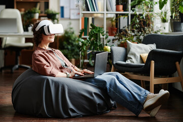 Leinwandbilder - Young modern businesswoman in vr headset and laptop on her knees sitting on bag chair and preparing presentation of new project