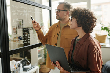 Poster - Young woman with laptop looking at notes on transparent board and listening to colleague explaining them during presentation
