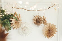 Stylish Paper Christmas Stars And Lights Hanging On White Wall Background. Scandinavian Festive Decoration In Boho Room. Handmade Paper Swedish Stars And Garland. Atmospheric Christmas Time
