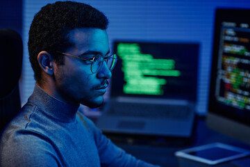 Leinwandbilder - Young serious IT support engineer in eyeglasses looking at computer screen while sitting in front of monitors and decoding data