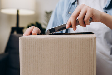 Wall Mural - Cropped shot of young female using utility knife to open parcel sitting on comfy couch at table. Close up woman received long-awaited box with gift. Delivery, mail, lifestyle and people concept