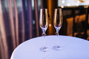 A pair of empty glass wine glasses stand on a round table with a white tablecloth at a wedding ceremony.