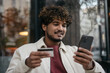 Smiling handsome Indian man holding credit card using mobile phone, shopping online outdoors, selective focus. Technology concept