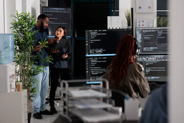 Two app developers analyzing html code looking for errors on laptop computer, checking big tv monitor in it agency office. Coders working on cyber security innovation in busy software company.