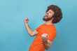 Side view of excited positive happy man with Afro hairstyle wearing orange T-shirt screaming for joy with raised fists and closed eyes, rejoicing win. Indoor studio shot isolated on blue background.