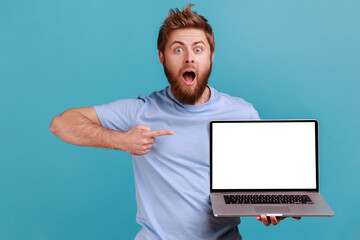 Wall Mural - portrait of shocked bearded man holding laptop and pointing at empty screen, being astonished with c