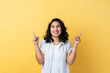 © khosrork - Portrait of satisfied delighted woman with dark wavy hair pointing up at empty place for ad content and expressing positive emotions. Indoor studio shot isolated on yellow background.
