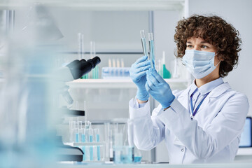 young female virologist in protective workwear looking at two flasks with liquid substances while co