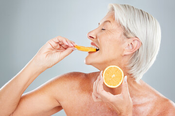 Beauty, orange and eating with a mature woman biting into a fruit slice in studio on a gray background. Food, wellness and natural care with a senior female posing to promote healthy eating or diet