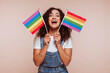 Smiling fascinating young arabian woman 20s years old wear white t-shirt and overalls, hold in hand striped colorful rainbow flags looking camera isolated over beige background studio portrait.