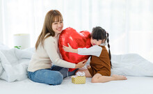 Millennial Asian Young Beautiful Mother Sitting On Bed Smiling Holding Hugging Red Heart Shaped Helium Air Balloon And Present Box With Little Cute Preschooler Daughter Girl In Bedroom In Morning