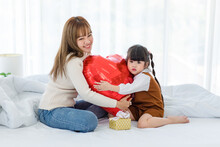 Millennial Asian Young Beautiful Mother Sitting On Bed Smiling Holding Hugging Red Heart Shaped Helium Air Balloon And Present Box With Little Cute Preschooler Daughter Girl In Bedroom In Morning