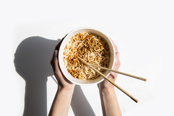 Female hands hold Chinese noodles in a cardboard bowl on a white background. Top view, fly lay