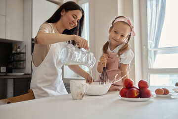 Wall Mural - An image of a young woman and her little cute daughter are cooking in the kitchen. Have fun together, interfere with flour. Mom teaches child to cook