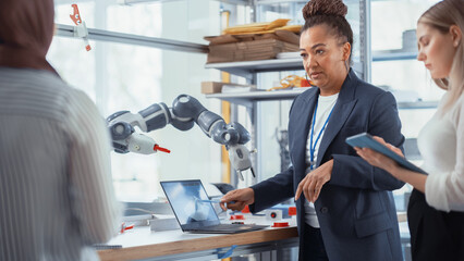Black Senior Engineer Explaining Information to People That Look at Robot Limb. Interns Hold Devices and Discussing. Optimisation of Autonomous Manufacturing Facility that Creates High Tech Products.