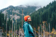 Lady with an orange hat stands in front of mountains with green fir trees.