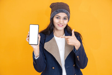 Poster - Happy young Indian girl wears blue winter jacket knitted hat showing smart phone with blank display screen to put advertisement or promotion isolated on orange yellow background studio portrait