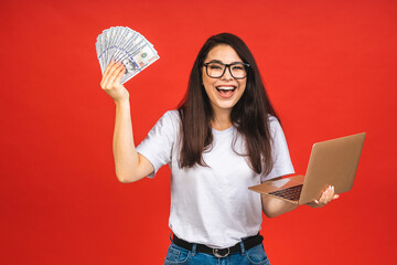 Canvas Print - Happy winner! Pretty young business woman in casual holding laptop in the office, isolated over red background. Working with computer, holding money bills.