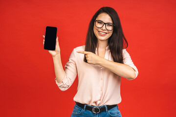 Canvas Print - Surprised happy smiling brunette woman showing blank smartphone screen and pointing on it while looking at the camera with smile isolated over red background.