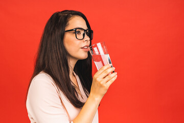 Wall Mural - Casual style young woman posing over isolated red studio background, holding a glass of water. Beautiful portrait of a girl. Female pose model. Healthy concept photo.