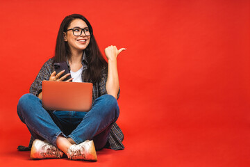 Canvas Print - Business concept. Portrait of happy smiling brunette woman in casual sitting on floor in lotus pose and holding laptop isolated over red background. Using mobile phone.