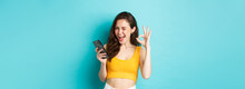 Technology And Lifestyle Concept. Cheerful Brunette Female Model Say Yes, Winking And Showing Okay Sign, Holding Mobile Phone, Standing Over Blue Background