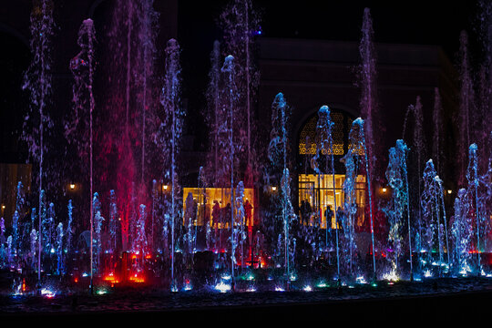 show of multi-colored musical fountains in siren tones at night Sirik against shops, horizontal