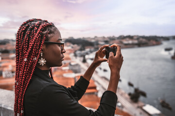 A profile shot of a young African female traveler with red box braids shooting a tourist attractions with her smartphone; a black tourist woman photographing urban landscape panorama via the cellphone