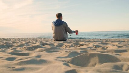 Wall Mural - Man on sandy beach drinking coffee from paper cup. Person on summer vacation relaxing and enjoying sea sunset. 4k video