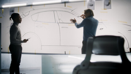 Two diverse automotive male designers discussing and developing futuristic electric car design. One drawing sketches on whiteboards, other holding tablet computer. Car design development studio.