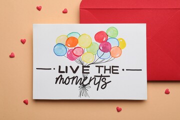 Wall Mural - Card with phrase Live The Moments and red envelope on beige background, flat lay