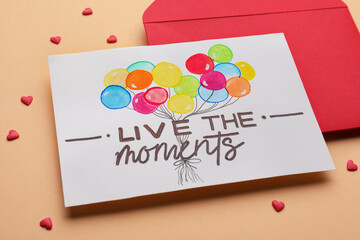 Wall Mural - Card with phrase Live The Moments and red envelope on beige background, closeup